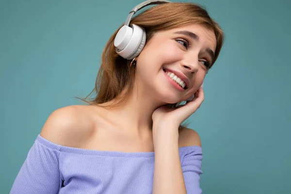 Closeup photo of pretty happy smiling young blonde woman wearing blue crop top isolated over blue background wall wearing white wireless bluetooth earphones listening to cool music and enjoying