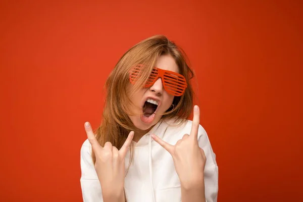 Tiro de bela jovem loira feliz emocional isolado sobre fundo laranja vestindo casuais capuz branco e óculos de sol elegantes mostrando gesto de rock and roll e se divertindo — Fotografia de Stock