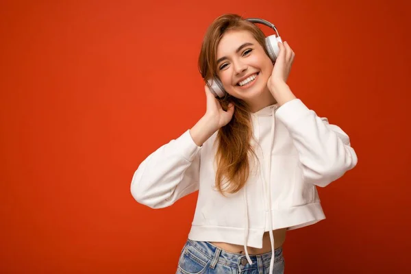 Photo of beautiful happy smiling young blonde woman wearing white hoodie isolated over colourful background wall wearing white wireless bluetooth earphones listening to cool music and enjoying looking — Stock Photo, Image