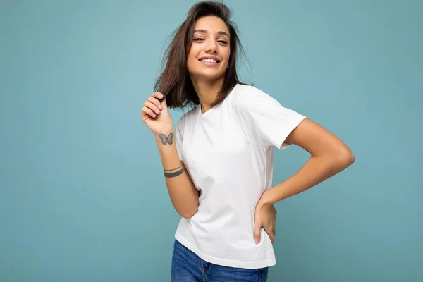 Портрет молодой красивой счастливой улыбающейся брюнетки в модной белой футболке с пустым местом для макетов. Сексуальная женщина, позирующая рядом с голубой стеной в студии с свободным пространством — стоковое фото