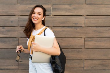 Güzel, gülümseyen, genç bir esmer kadın elinde bilgisayar dizüstü bilgisayar, beyaz tişörtlü güneş gözlüğü ve sokakta açık mavi kot pantolon tutuyor.