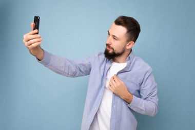 Yakışıklı, şık kıyafetler giyen arka plandaki duvarda izole bir şekilde duran akıllı telefon taşıyan cep telefonu ekranına bakarak selfie çeken genç bir adam.