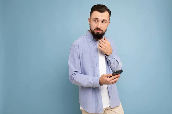 Pensativo joven guapo aislado sobre la pared de fondo usando ropa cotidiana sosteniendo y usando sms de escritura de teléfonos móviles mirando a un lado y pensando — Foto de Stock