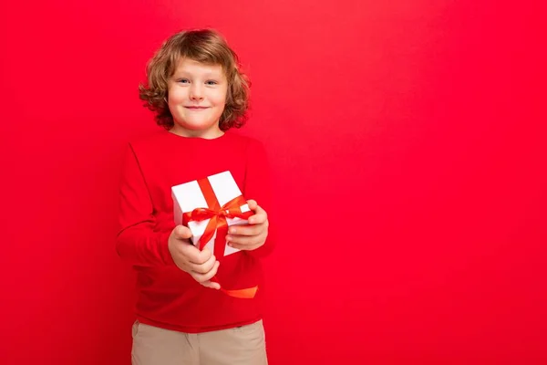 Foto de alegre sonriente rubia rizado chico aislado sobre fondo rojo pared usando suéter rojo celebración de caja de regalo y mirando a la cámara — Foto de Stock