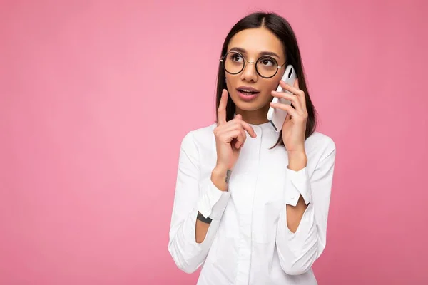Vacker ung brunett kvinna bär vit blus och optiska glasögon står isolerade över rosa bakgrund talar på mobiltelefon tittar åt sidan och tänker — Stockfoto
