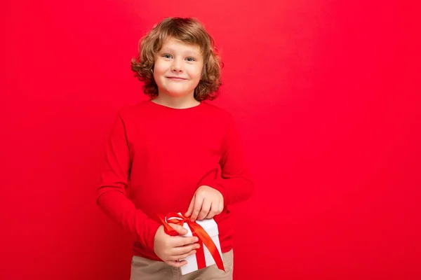 Positivo sonriente rubia rizado chico aislado sobre fondo rojo pared usando suéter rojo celebración de caja de regalo y mirando a la cámara — Foto de Stock