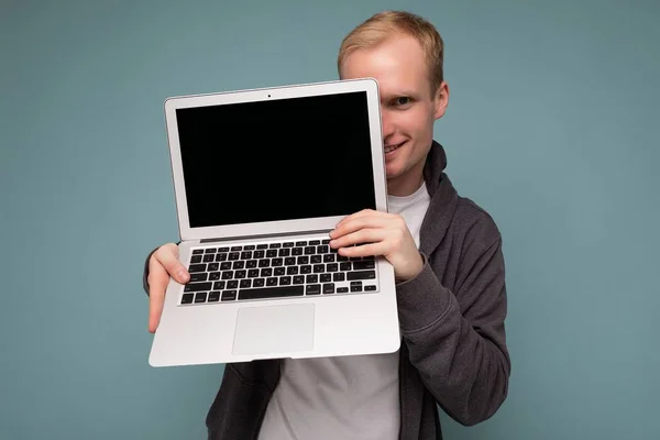 Foto de un guapo hombre rubio sonriente sosteniendo un ordenador portátil con pantalla de monitor vacía con maqueta y espacio de copia con camiseta blanca y suéter gris mirando a la cámara aislada sobre azul —  Fotos de Stock