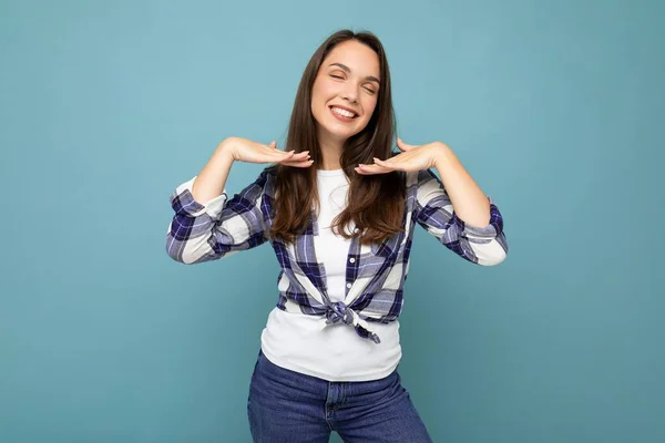 Снимок молодой позитивной восхитительной улыбающейся красивой брюнетки с искренними эмоциями в модной чековой рубашке, стоящей изолированно на синем фоне с бесплатным пространством — стоковое фото