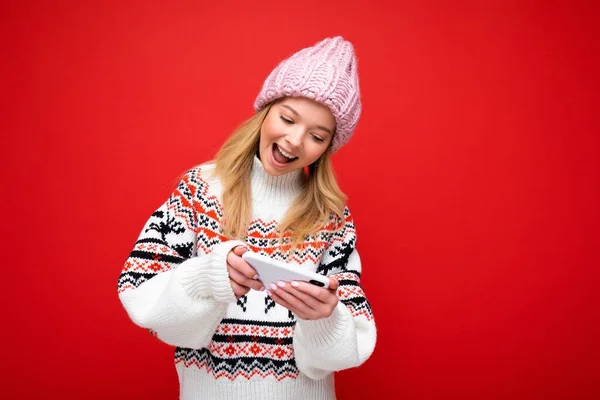 Foto av vackra leende ung blond kvinna bär varm stickad hatt och vinter varm tröja stående isolerad över röd bakgrund spela spel via smartphone tittar på enhet display — Stockfoto