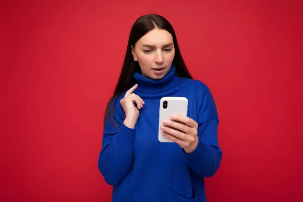 Triste atraente boa aparência jovem morena vestindo elegante camisola quente azul balançando isolado no fundo vermelho com espaço vazio segurando na mão e usando sms de mensagens de telefone móvel olhando — Fotografia de Stock