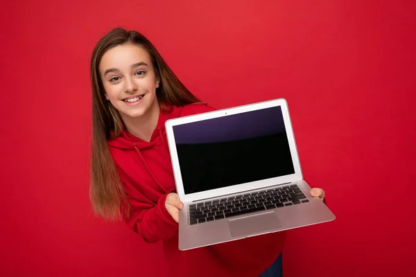 Foto de retrato de cerca de la hermosa chica sonriente feliz con el pelo largo con capucha roja que sostiene el ordenador portátil mirando a la cámara aislada sobre el fondo de la pared roja —  Fotos de Stock