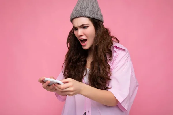 魅力的な感情的な若いブルネットの女性の写真スタイリッシュなピンクのシャツとグレーの帽子を身に着けているピンクの背景を手に持っている上に隔離され、携帯電話を使用してオンラインゲームをプレイするgadjetを見て — ストック写真