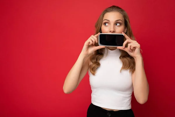 Funny attractive young blonde female person wearing white t-shirt isolated on red background with copy space holding smartphone showing phone in hand with empty screen for cutout looking to the side — Stock Photo, Image