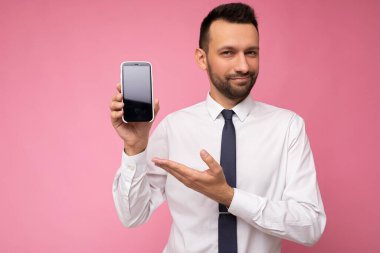 Yakışıklı, yakışıklı, yakışıklı, şık kıyafetler giyen, renkli arka plan duvarında tek başına duran, akıllı telefon tutan ve boş ekran ekranlı telefonu gösteren yakışıklı bir adam.