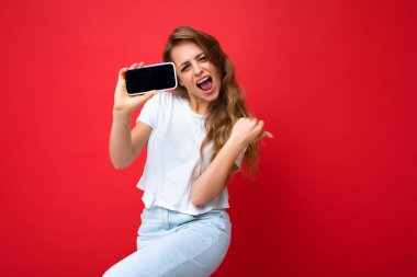 Mutlu, güzel, gülümseyen, güzel, sarışın, güzel, beyaz tişörtlü, kırmızı arka planda izole bir şekilde duran, elinde telefonuyla elinde akıllı telefonu boş ekranla elinde tutan güzel bir kadın.
