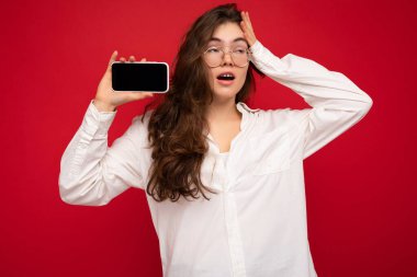 Kırmızı arka plan üzerinde izole edilmiş beyaz gömlek ve optik gözlük takan büyüleyici genç esmer kadın elinde cep telefonu ile yan tarafa bakıyor.
