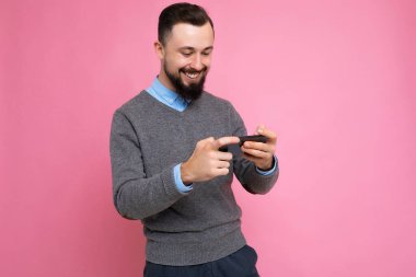 Arka plandaki duvarda izole edilmiş gri süveter ve mavi gömlek giyen, sakallı, mutlu, genç, esmer bir adamın cep telefonuyla oyun oynaması.