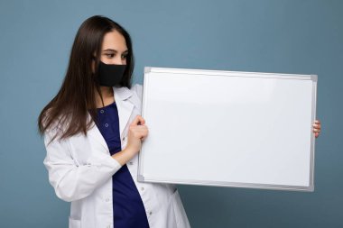 Çekici esmer kadın portresi beyaz ceketli ve siyah maskeli beyaz pano tutan mavi arka planda izole edilmiş beyaz pano.