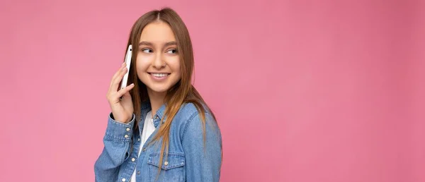 Панорамное фото привлекательной позитивной улыбающейся молодой блондинки в повседневной синей джинсовой рубашке, изолированной на розовом фоне, держащей в руке и разговаривающей по мобильному телефону, глядя в сторону — стоковое фото