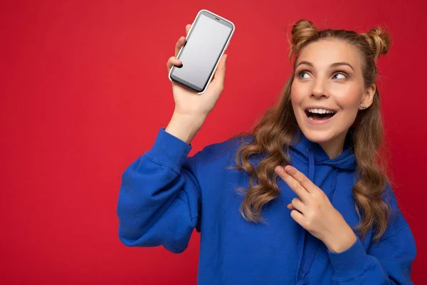 Aantrekkelijke glimlachende jonge blonde vrouw draagt stijlvolle blauwe hoodie geïsoleerd op rode achtergrond met kopieerruimte houden smartphone tonen telefoon in de hand met leeg scherm wijzen naar gadjet — Stockfoto