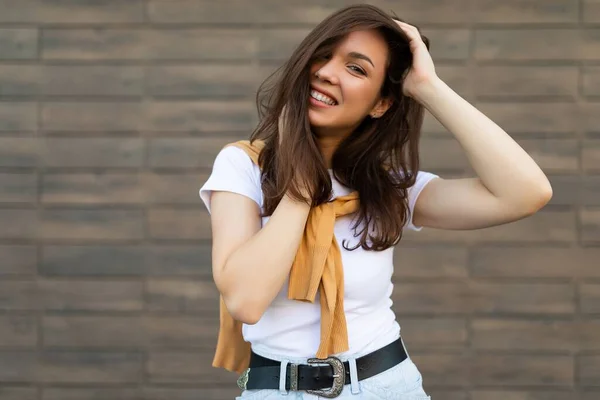 Портрет молодої красивої позитивної щасливої брюнетки, що стоїть біля коричневої стіни на вулиці і носить стильний одяг — стокове фото