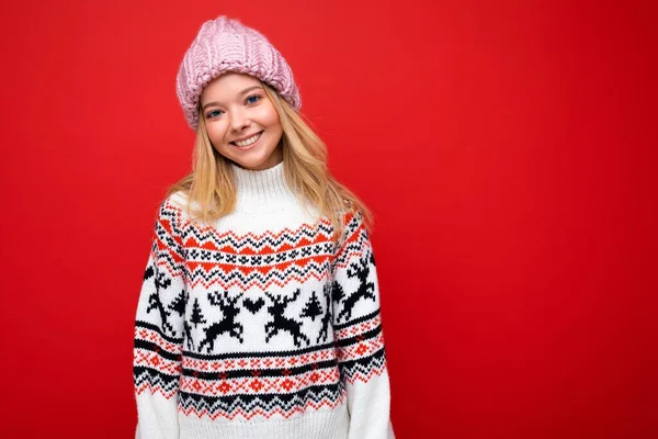 写真かなり肯定的な笑顔若いですブロンド女性絶縁以上赤の背景壁身に着けています冬のセーターとピンクの帽子カメラを見て楽しいです — ストック写真