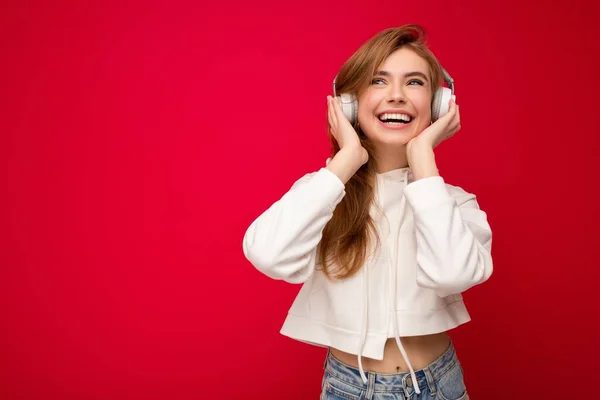 Foto de bela feliz sorrindo jovem loira vestindo capuz branco isolado sobre parede de fundo colorido vestindo fones de ouvido sem fio bluetooth branco ouvindo música legal e gostando de olhar — Fotografia de Stock