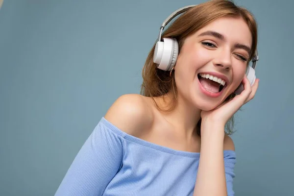 Foto de primer plano de la atractiva mujer rubia joven y sonriente que usa top azul de la cosecha aislado sobre la pared de fondo azul usando auriculares escuchando música y divirtiéndose — Foto de Stock