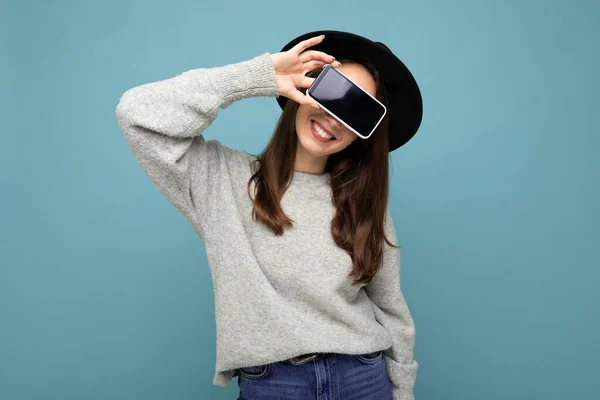 Mulher sorridente muito jovem usando chapéu preto e camisola cinza segurando telefone olhando para a câmera isolada no fundo — Fotografia de Stock