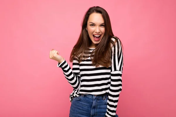 stock image Portrait of young positive happy attractive brunette woman with sincere emotions wearing casual striped pullover isolated on pink background with empty space and celebrating winning shouting yeah