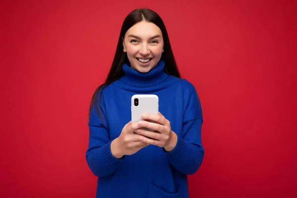 Lachende aantrekkelijke positief uitziende jonge brunette vrouw dragen stijlvolle blauwe warme trui evenwicht geïsoleerd op rode achtergrond met lege ruimte in de hand en met behulp van mobiele telefoon berichtgeving sms — Stockfoto