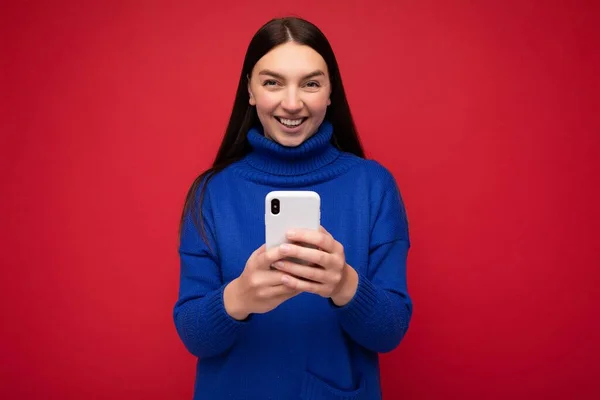 Sorrindo atraente positivo boa aparência jovem morena vestindo elegante camisola quente azul balançando isolado no fundo vermelho com espaço vazio segurando na mão e usando sms de mensagens de telefone móvel — Fotografia de Stock