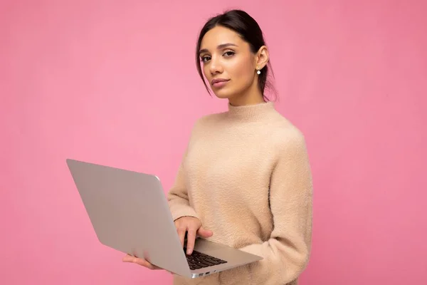 Fotografie de frumos serios tânără brunetă femeie purtând pulover bej care deține netbook calculator tastarea text pe tastatură uitându-se la camera izolată peste fundal de perete roz — Fotografie, imagine de stoc