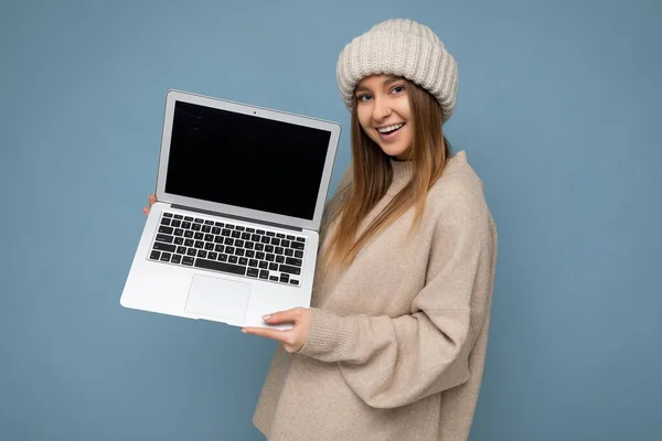 Frumos zâmbind fericit tânără blondă întunecată femeie în iarnă pălărie călduroasă tricotată ținând laptop-ul computerului uitându-se la aparatul foto purtând pulover de iarnă bej izolat peste fundal de perete albastru deschis în studio — Fotografie, imagine de stoc