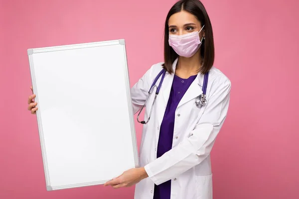 戴防护面罩，身穿白色医疗服，手持用粉色背景隔离的空磁板的护士 — 图库照片