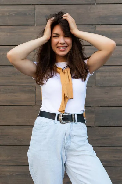 Vertical ptoto of young beautiful positive happy brunette woman standing against brown wall in the street and wearing stylish outfit — Stock Photo, Image