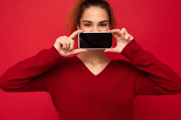 Close-up Foto van vrolijke grappige jonge vrouw met donkerrode trui geïsoleerd over rode achtergrond holding smartphone en toont mobiele telefoon scherm met kopieerruimte voor cutout kijken naar camera — Stockfoto