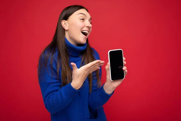 Photo of pretty happy young brunette woman wearing casual blue sweater isolated over red background with empty space holding in hand mobile phone and showing smartphone with empty screen for mockup — Stock Photo, Image