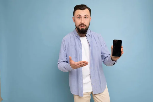 Foto de pedir chateado bonito boa aparência jovem morena homem sem barba com barba vestindo t-shirt branca casual e camisa azul balançando isolado no fundo azul com espaço vazio segurando na mão — Fotografia de Stock