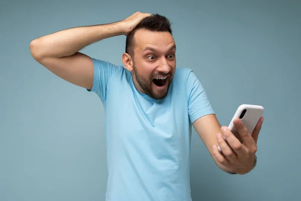 Потрясенный эмоциональный красивый молодой бородатый брюнетка мужчина в повседневной синей футболке изолированы на синем фоне холдинг и использование мобильной связи в Интернете глядя на gadjet — стоковое фото