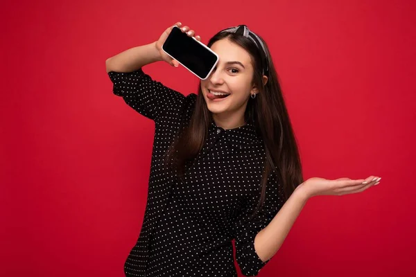 Delicioso sorriso Menina bonita morena de pé isolado sobre a parede vermelha vestindo roupas pretas elegantes casuais mostrando telefone celular com tela vazia para recorte olhando para a câmera — Fotografia de Stock