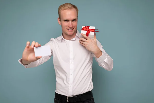 Foto de guapo feliz sonriente joven hombre rubio aislado sobre la pared de fondo azul con camisa blanca sosteniendo caja de regalo blanca con cinta roja y tarjeta de crédito mirando a la cámara —  Fotos de Stock