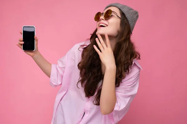 Close-up foto van Positieve aantrekkelijke jonge brunette vrouw dragen stijlvolle roze shirt grijze hoed en kleurrijke zonnebril geïsoleerd over roze achtergrond vasthouden in de hand en toont mobiele telefoon met lege — Stockfoto