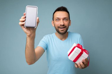 Yakışıklı, pozitif esmer, sakallı, mavi arka plan duvarında yaşayan, mavi tişörtlü, kırmızı kurdeleli beyaz hediye kutusu ve boş yeşil ekranlı cep telefonu olan genç bir adam.