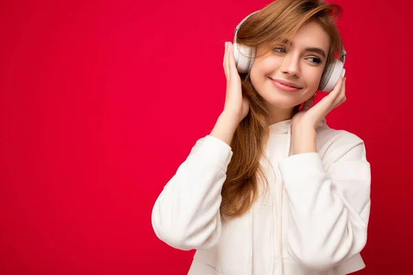 Photo of beautiful happy smiling young blonde woman wearing white hoodie isolated over colourful background wall wearing white wireless bluetooth earphones listening to cool music and enjoying looking — Stock Photo, Image