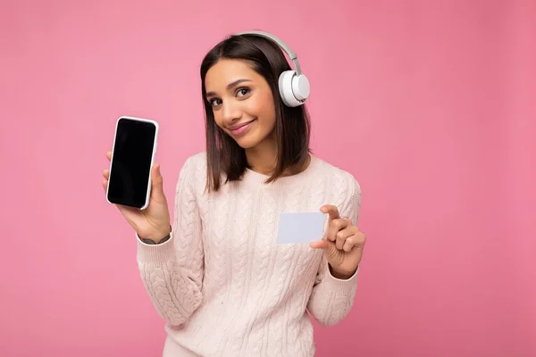 핑크 색 바탕에 고립된 핑크 색 스웨터를 입고 있는 아름다운 젊은 갈색 머리 여인은 무선 헤드폰을 쓰고 음악을 듣고 빈 핸드폰을 보여 주고 있다. — 스톡 사진