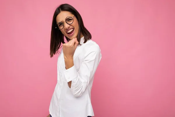 Jovem positivo feliz legal bela morena mulher vestindo camisa branca e óculos ópticos isolados sobre fundo rosa com espaço vazio e mostrando gesto de rock and roll — Fotografia de Stock