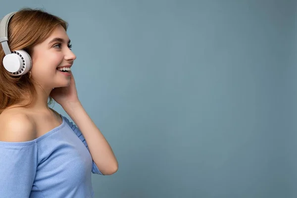Side-profile photo of charming positive smiling young blonde woman wearing blue crop top isolated over blue background wall wearing white wireless bluetooth headphones listening to cool music and — Stock Photo, Image