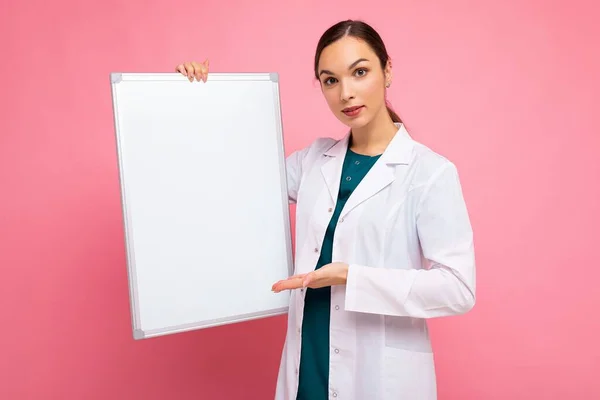 Ritratto di attraente giovane donna bruna positiva che indossa un cappotto bianco medico con bordo magnetico bianco per il modello isolato su sfondo rosa con spazio per la copia — Foto Stock