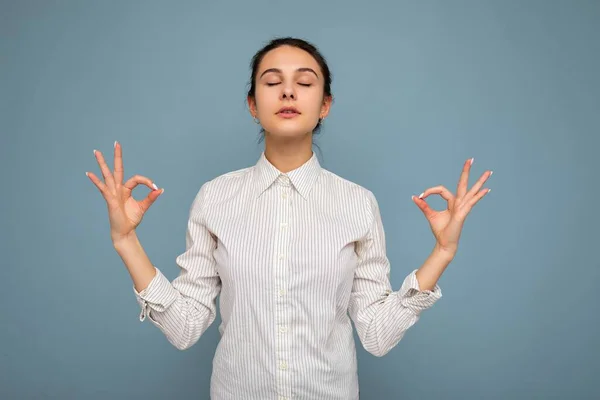 Foto de una joven morena concentrada con emociones sinceras usando una camisa blanca aislada sobre un fondo azul con espacio vacío y riendo y manteniendo la mano en señal de mudra. Conjunto de yoga — Foto de Stock